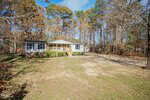 A single-story house with a small front porch and steps, white siding, and green shutters, sits on a spacious lot with a dirt driveway, surrounded by trees with autumn foliage.