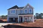 A two-story unfinished house with gray siding and white trim, featuring a covered front porch, double garage, construction materials on site, and red dirt piles in the yard against a clear blue sky.
