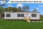 A single-story, light-colored manufactured home with black shutters and a small front porch sits on a grassy lot with landscaping. A banner at the top reads, "New Construction on 1+ Acre Lot." Trees are in the background.