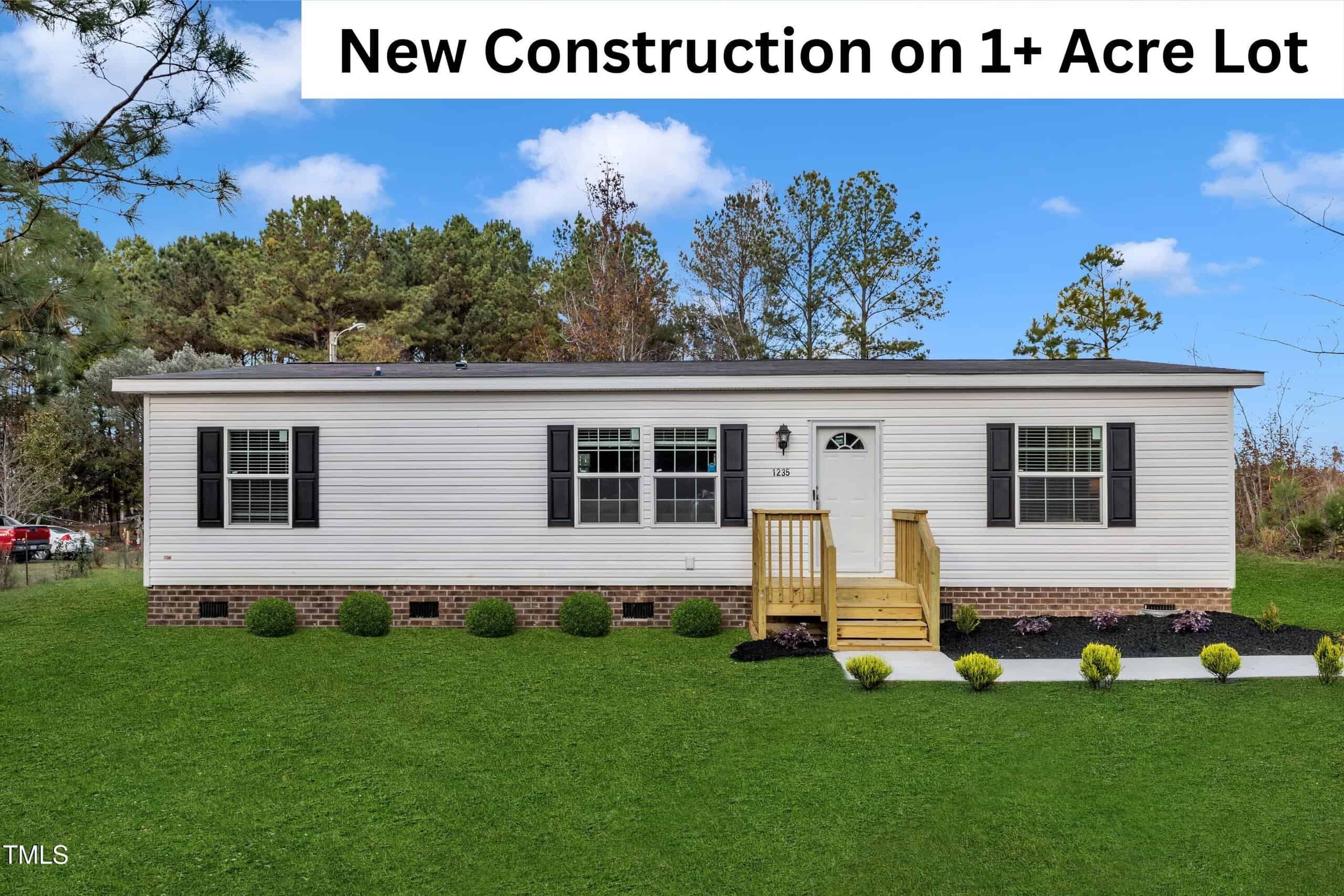 A single-story, light-colored manufactured home with black shutters and a small front porch sits on a grassy lot with landscaping. A banner at the top reads, "New Construction on 1+ Acre Lot." Trees are in the background.