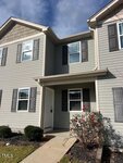 Two-story gray townhouse with white trim and dark shutters. There are three front windows and a white entry door. Small shrubs and flowering bushes line the short walkway leading to the entrance.