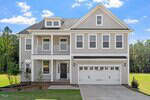 Two-story light gray house with white trim, a covered front porch, double garage, and five front-facing windows. The house sits on a lawn with minimal landscaping and is bordered by trees in the background.