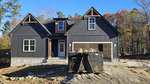 A newly constructed dark gray house with white trim and a stone foundation, featuring a front porch, a double garage, and a large dumpster filled with construction debris in the driveway. Trees with autumn leaves are in the background.
