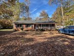 A single-story brick house with a covered front porch and black shutters sits amid autumn trees. Fallen leaves cover the front yard, and a blue SUV is parked to the right of the house.