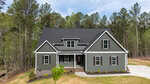 A two-story house with dark gray siding and white trim, a covered front porch, and a black shingled roof. The home is surrounded by tall trees, with a concrete driveway and minimal landscaping in front.
