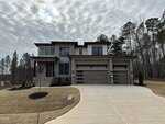 A modern two-story house with light and dark exterior finishes, large windows, a three-car garage, and a wide driveway, surrounded by bare trees and landscaped yard under a cloudy sky.