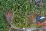 Aerial view of a triangular, wooded lot outlined in yellow, located on a corner next to a paved road. A single house with a driveway is visible to the right, and several cars are parked along the road.