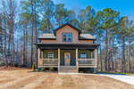 A two-story house with tan siding, a covered front porch, and two dormer windows, set among tall pine trees with a dry lawn and a concrete driveway on a clear day.