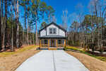 A two-story modern house with white and gray exterior, black trim, and a covered front porch is situated at the end of a long concrete driveway surrounded by trees and brown grass.