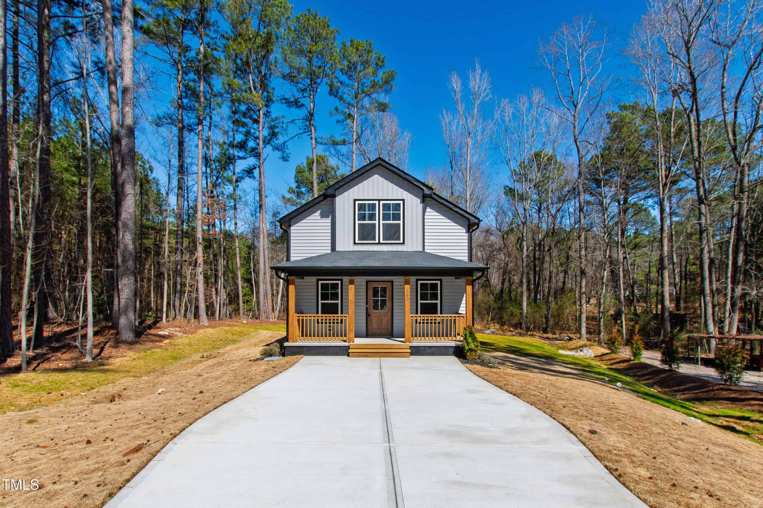 A two-story modern house with white and gray exterior, black trim, and a covered front porch is situated at the end of a long concrete driveway surrounded by trees and brown grass.