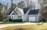 A gray, single-story house with white trim, a one-car garage, and a basketball hoop in the driveway. The yard is grassy with trimmed bushes along the front and trees in the background.