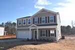 A two-story suburban house with beige siding, white trim, black shutters, and a covered entry porch. The home has a two-car garage and sits on a newly landscaped lawn with a small tree in the front yard.