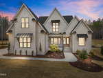 A modern two-story house with light-colored siding and brick, black window trim, multiple gables, covered entryway, landscaped front yard, and a driveway, pictured at dusk with surrounding trees in the background.