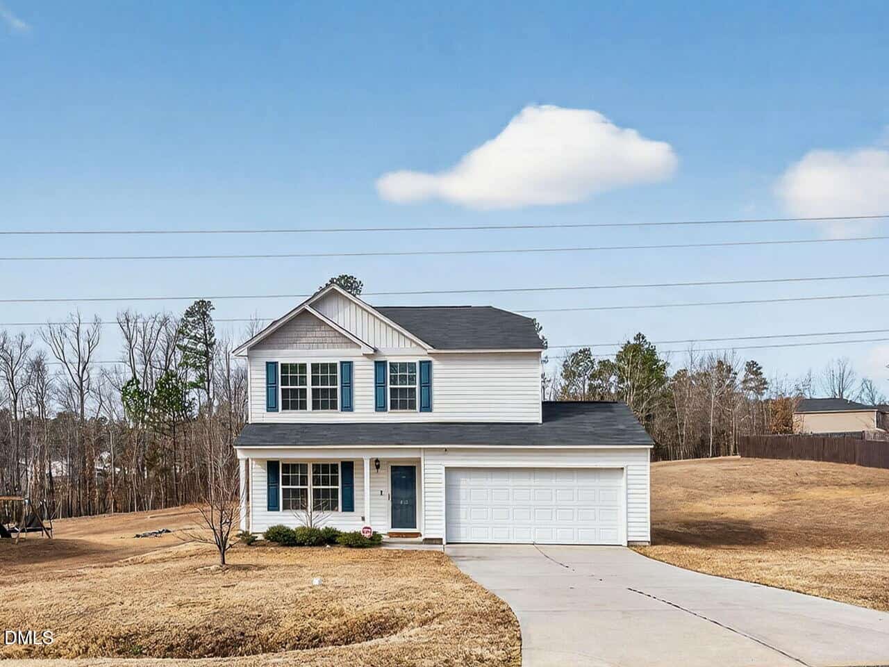 A two-story white house with black shutters and a covered front porch. The home has a double garage and sits on a large, grassy lot with few trees and clear blue sky in the background.