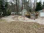 A backyard with a raised wooden deck, a small shed, a white garage, and scattered gravel and leaves. There are trees without leaves in the background and an American flag attached to the garage.