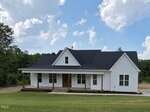 A white, single-story farmhouse with a black roof, wooden porch columns, and a covered front porch sits on a grassy lawn with trees in the background under a blue sky with scattered clouds.