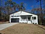A modern single-story house with white brick exterior, black trim, and a black double garage. The home has a front porch with steps and sits on a newly sodded lawn surrounded by tall trees.