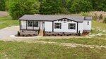 A single-story manufactured home with a covered front porch, white exterior, dark trim, and a stone foundation, situated on a grassy lot with trees and shrubs in the background.