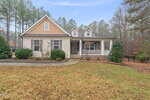 Single-story house with beige siding, stone accents, and a covered front porch, set on a grassy yard with shrubs and bordered by tall pine trees. A curved walkway leads to the front door.