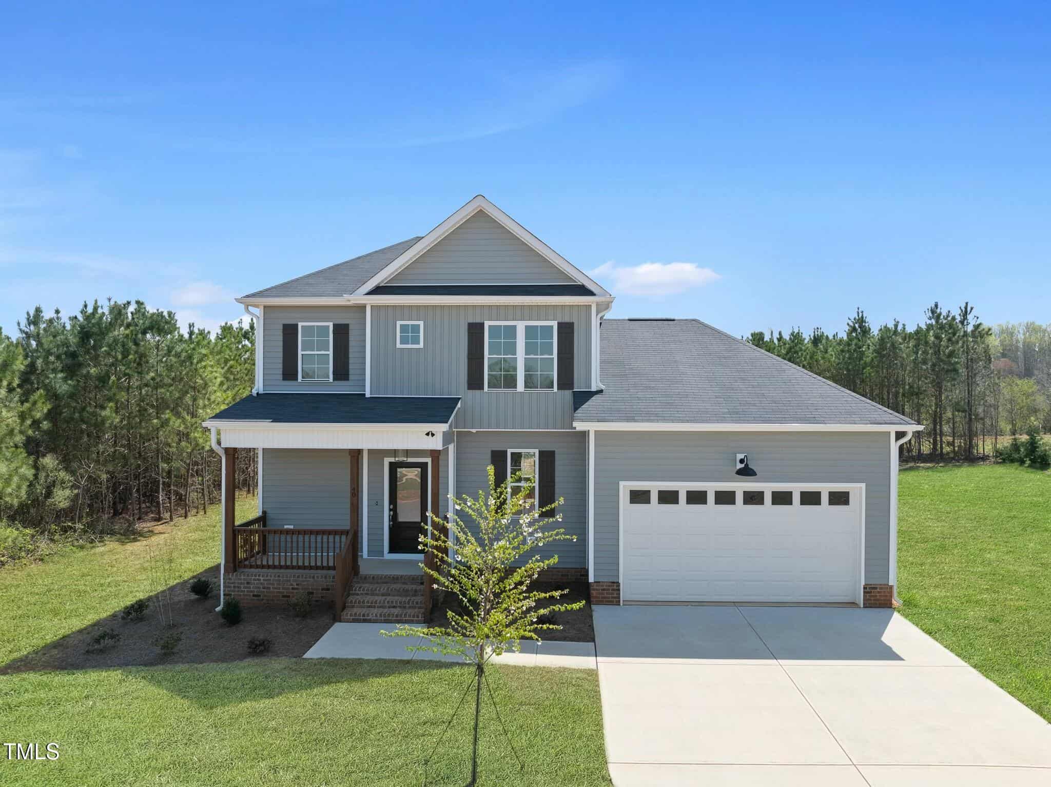 A two-story gray house with white trim, a covered front porch, and an attached two-car garage, situated on a large green lot bordered by trees under a clear blue sky.