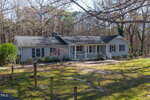 Single-story light gray house with a covered front porch, white railings, and dark shutters, surrounded by trees and a grassy yard with some bare patches. The roof shows signs of age.