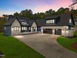 A modern two-story house with gray stone siding and dark accents, featuring three garage doors and large windows. The driveway is wide and curved, and the surrounding yard is neatly landscaped with green grass.