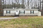 A single-story manufactured home with beige and white siding, black shutters, a small front porch with steps, and a gravel driveway, surrounded by a grassy yard and bare trees.