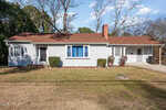 Single-story house with light gray siding, brown shingle roof, and blue curtains in the front window. There is a small front yard with sparse grass, shrubs by the house, and a driveway on the right side.