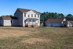 Two-story beige house with a covered front porch, detached three-car garage, and an RV parked nearby. The yard is spacious with patchy grass, and there are trees in the background under a clear blue sky.