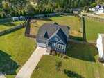 Aerial view of a single-family house with a gray roof, attached garage, and front porch, situated on a large, fenced, grassy lot with trees and a small shed in the backyard. Driveway leads to the street.
