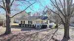 A small, single-story yellow house with white trim and a front porch sits in a yard with bare trees and sparse grass. There are hedges along the front of the house and a white shed visible in the background.