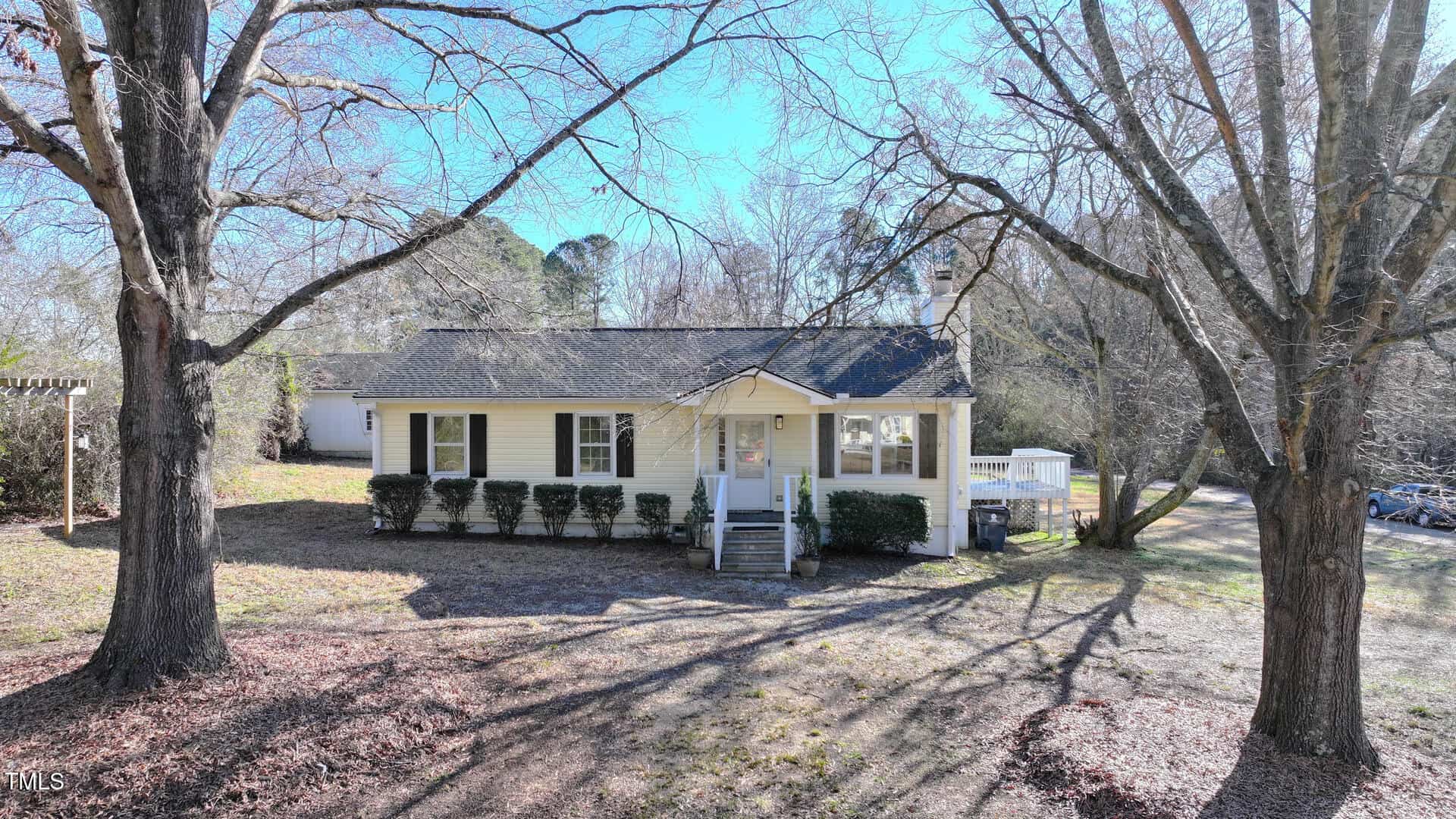 A small, single-story yellow house with white trim and a front porch sits in a yard with bare trees and sparse grass. There are hedges along the front of the house and a white shed visible in the background.