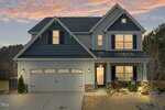 Two-story house with dark blue shingles and light gray siding, illuminated windows, a double garage, small front porch with two chairs, and a landscaped yard at sunset.