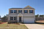 Two-story suburban house with beige siding, black shutters, a double garage, and a small front lawn. The home is set against a backdrop of trees and an unfinished yard.