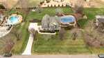 Aerial view of a two-story house with a gray roof, large driveway, landscaped front yard, fenced backyard, and an in-ground pool covered with a blue tarp. Surrounding areas have open grassy spaces and neighboring homes.