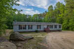 A single-story manufactured home with gray siding and black shutters sits on a large dirt lot, surrounded by trees and large rocks under a partly cloudy sky. A small red staircase leads to the front door.