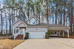 A two-story suburban house with gray siding, white trim, a front porch with brick steps, an attached two-car garage, a carport, and surrounded by trees on a concrete driveway.
