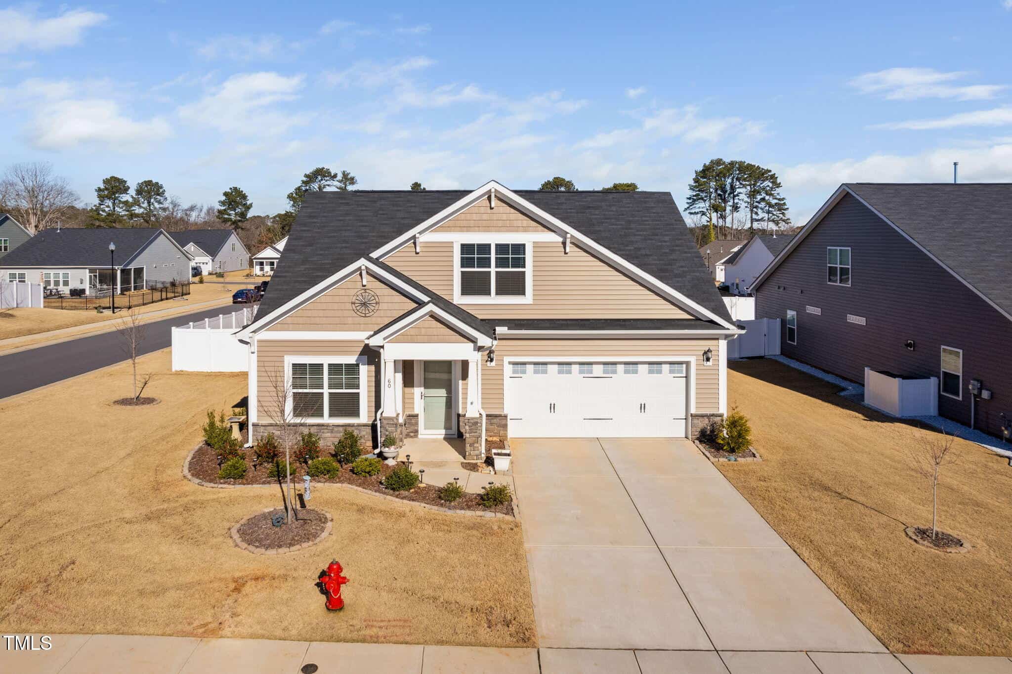 A single-story suburban house with beige siding, white trim, and a two-car garage sits on a large lot with a concrete driveway and minimal landscaping. A red fire hydrant is visible near the driveway.