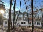 A white manufactured home with black shutters sits in a wooded area. Tall leafless trees surround the home, and sunlight filters through the branches. The ground is covered with dry leaves and patches of dirt.