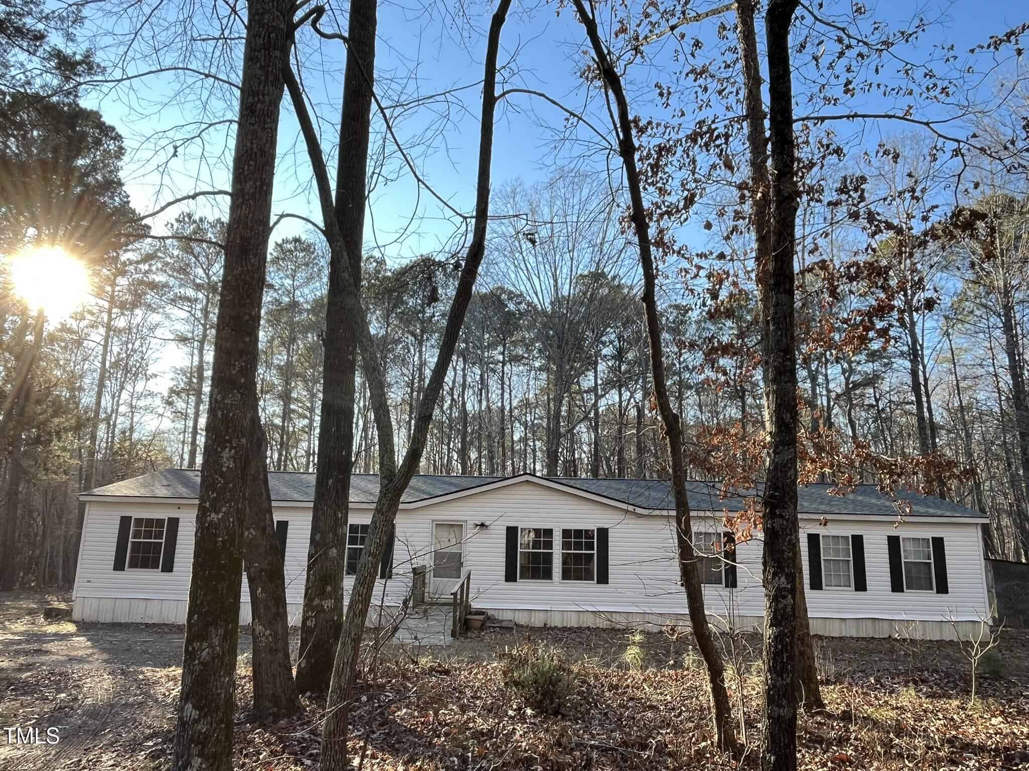 A white manufactured home with black shutters sits in a wooded area. Tall leafless trees surround the home, and sunlight filters through the branches. The ground is covered with dry leaves and patches of dirt.