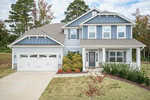 Two-story house with gray and blue siding, white trim, a front porch, double garage, and a neatly landscaped yard with shrubs and small trees. Pines and other trees are visible in the background.