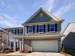 A two-story suburban house with tan siding, dark blue shutters, multiple windows, and a double garage. The home has a covered entryway and minimal landscaping, under a clear blue sky.