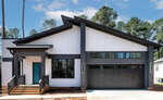 A modern single-story house with white vertical siding, black trim, a teal front door, and a black double garage. The driveway and porch are unfinished, and tall trees are visible in the background.