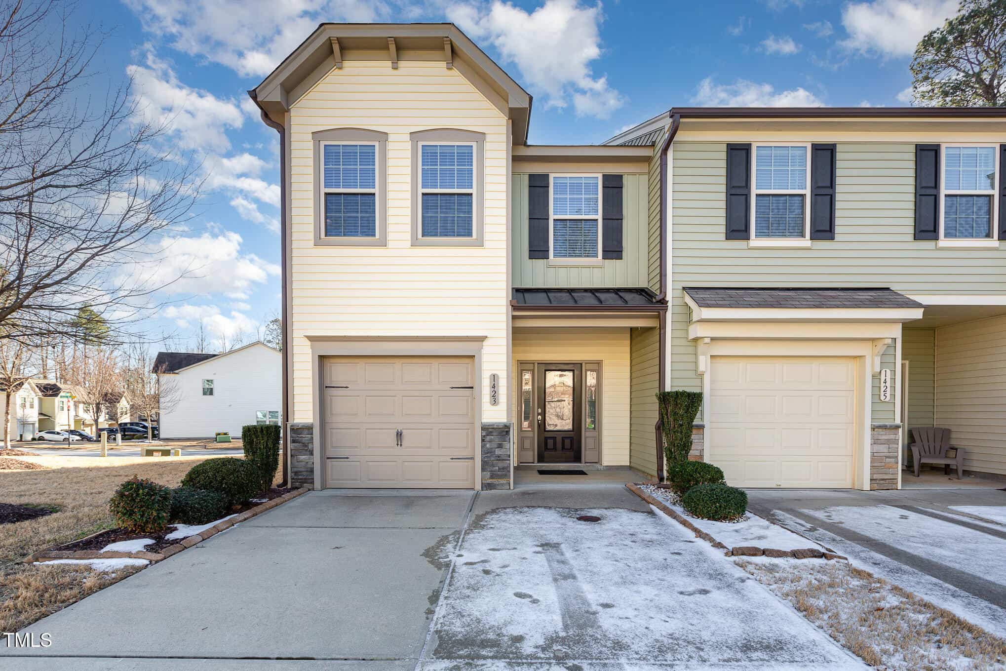 A two-story beige townhouse with a single-car garage, black shutters, and a small covered front entry. Patchy snow covers parts of the driveway and lawn. The sky is mostly clear with a few clouds.