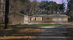 Single-story house with beige siding, white trim, and attached two-car garage. The house has a front porch with steps, exterior lighting, a star decoration, and is surrounded by tall trees and a curved driveway.