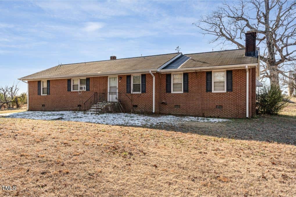A single-story red brick house with a gable roof, three visible chimneys, and a small front porch. There is patchy snow on the ground near the foundation and the yard is mostly brown grass. The sky is mostly clear.