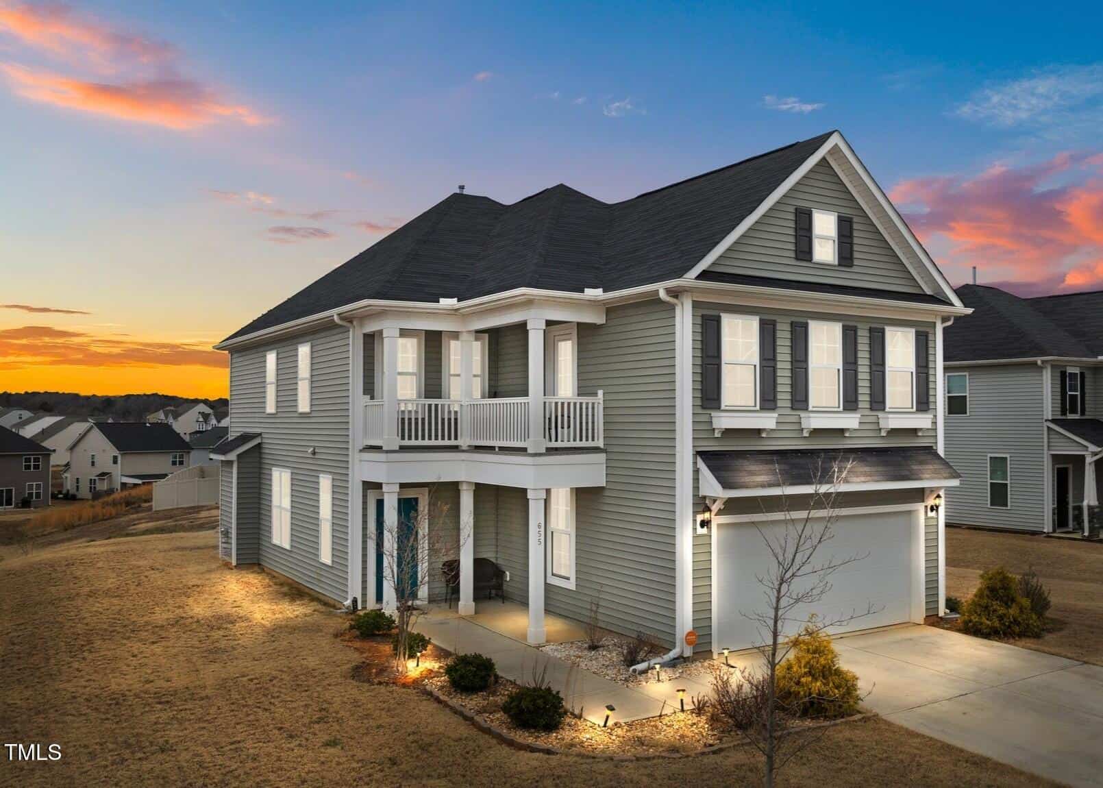 Two-story gray house with white trim, a covered porch, balcony, and attached two-car garage. The house is surrounded by a lawn and is situated in a suburban neighborhood at sunset.