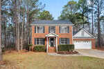 Two-story brick house with white trim, black shutters, and a front porch. Attached two-car garage on the right. Surrounded by tall pine trees and situated on a large, grassy lot with a concrete driveway.