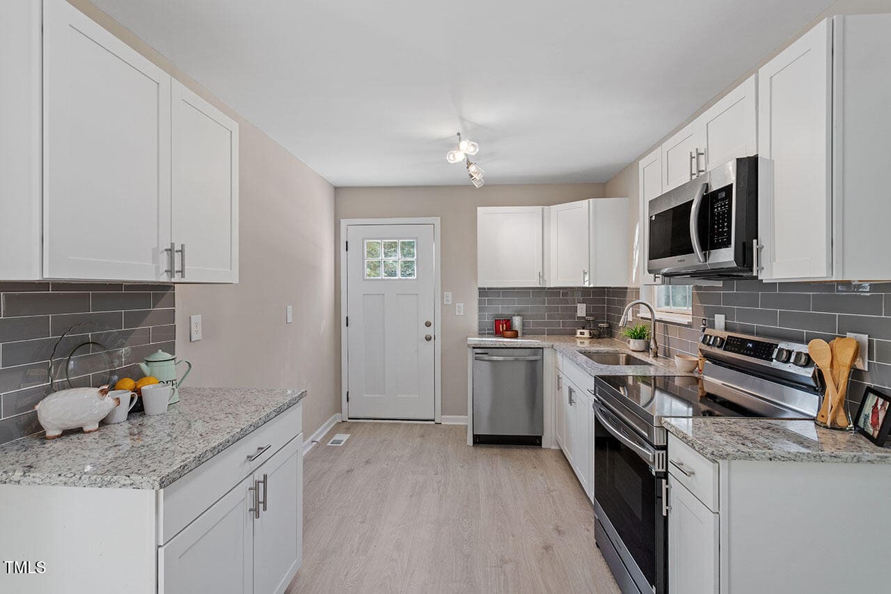 Modern kitchen with white cabinets, stainless steel appliances, gray subway tile backsplash, granite countertops, and light wood flooring. A door with a window is at the back, and small kitchen items are on the counters.
