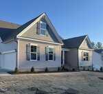 A single-story house with gray siding, white trim, and a two-car garage. The yard has fresh sod and small shrubs planted near the foundation. The sky is clear and blue.